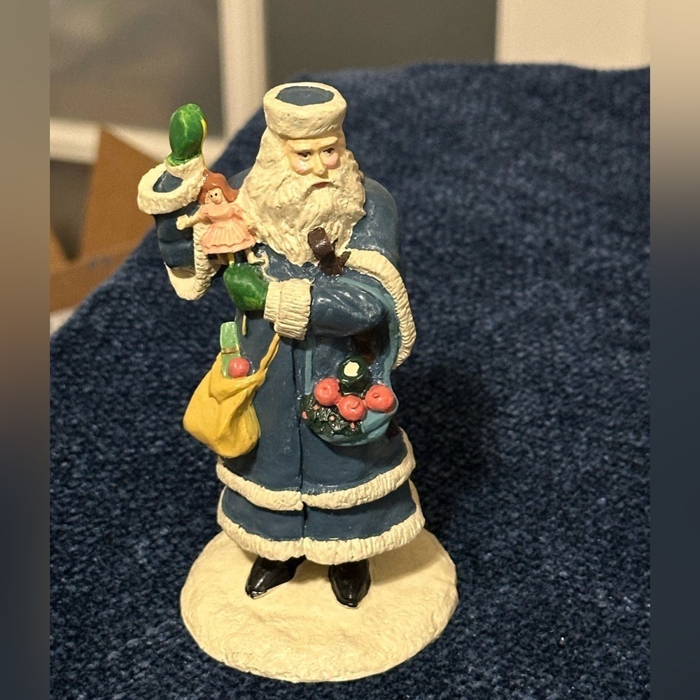 Blue robed Santa with green mittens holds a doll & basket of fruit.